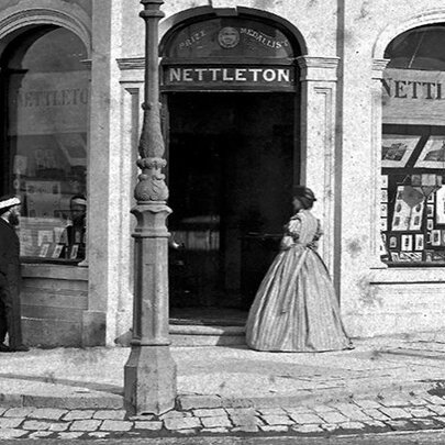 A black and white photograph, circa 1870, of a corner shop at 1 Madeline St, Carlton, showing a woman standing at the entrance and two men looking in the windows.