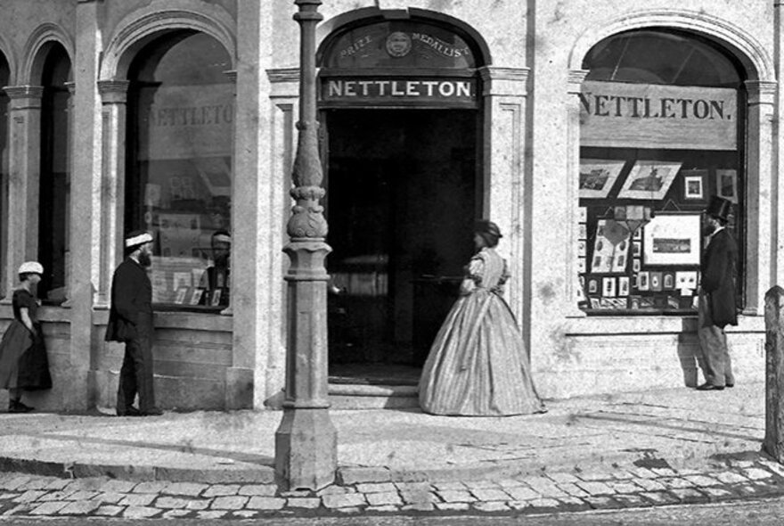 A black and white photograph, circa 1870, of a corner shop at 1 Madeline St, Carlton, showing a woman standing at the entrance and two men looking in the windows.