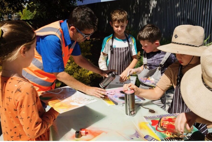 A man wearing a high-vis vest, showing a group of children how to use spray paint to create art.