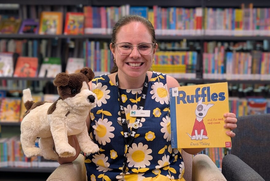 A woman holding a picture book titled 'Ruffles' and also holding a dog toy in a library reading area. 