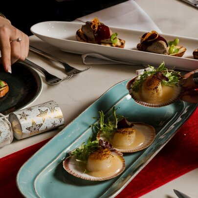A festive dining table featuring a selection of seafood (such as seared scallops on half-shells) and small bites served on decorative platters with a Christmas bob-bon cracker on the table.
