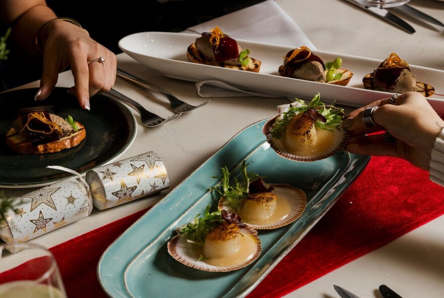 A festive dining table featuring a selection of seafood (such as seared scallops on half-shells) and small bites served on decorative platters with a Christmas bob-bon cracker on the table.
