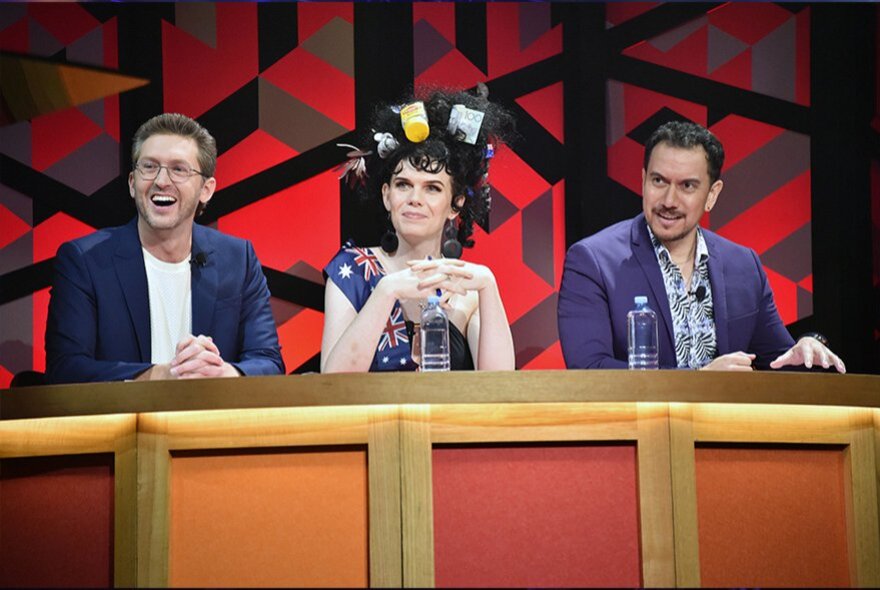 A trio of comedians seated behind a desk on a stage as part of the The Annual Great Debate.