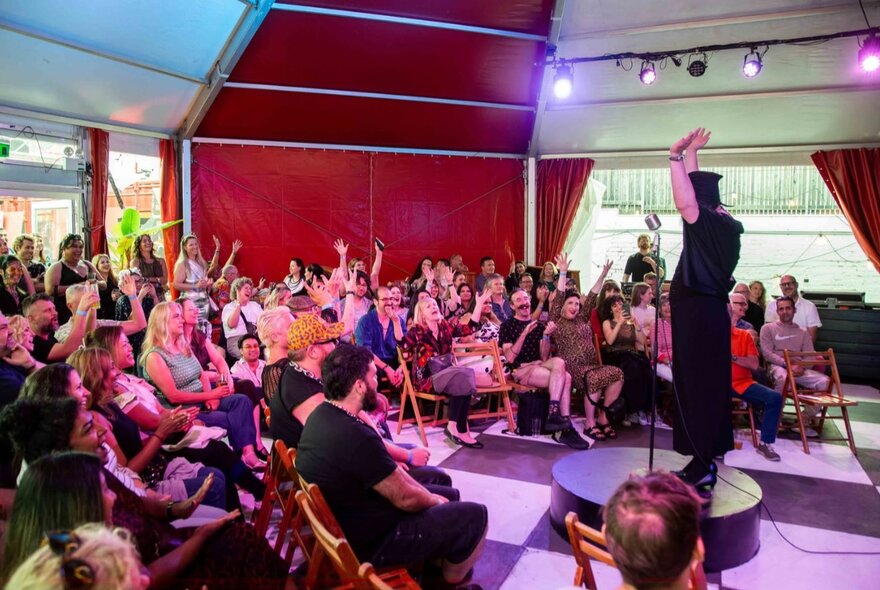 A woman on a round pedestal with her hands in the air, a circular audience watching on.