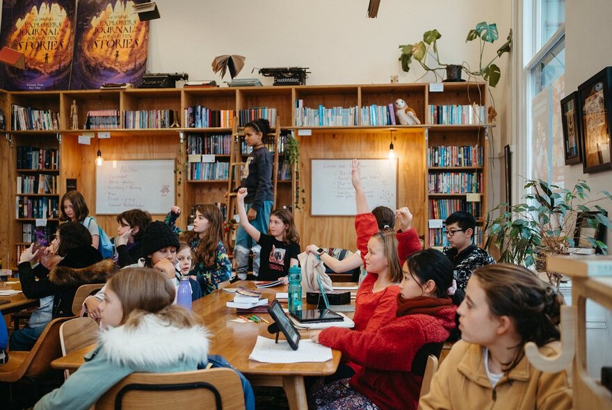 Teenagers sitting and working around a large table in a room, some with laptops open in front of them, bookshelves in the background.