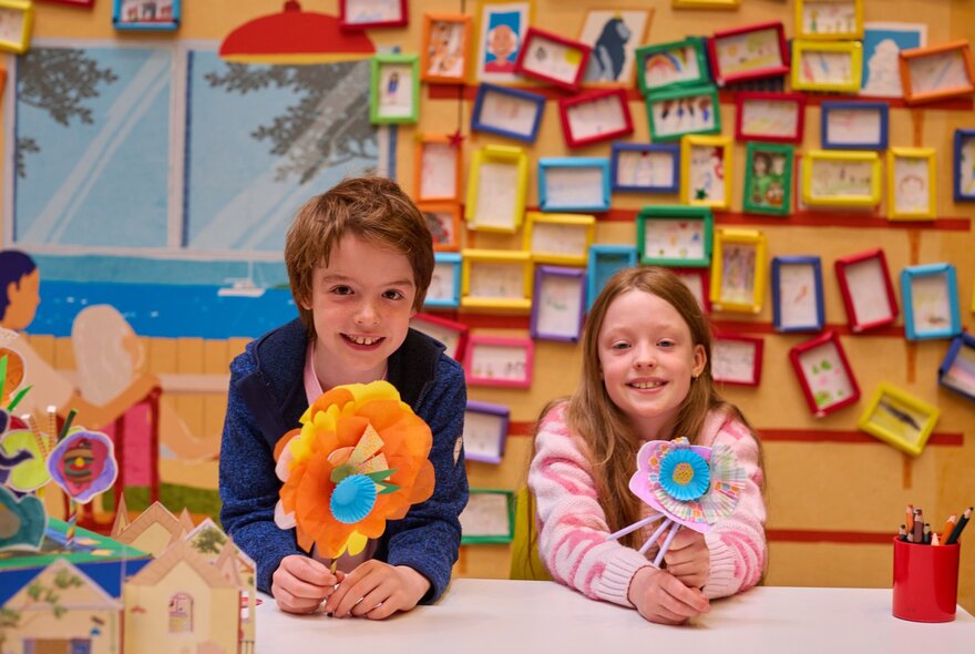 Two children proudly showing their handcrafted paper flowers in a colouful room with tens of coloured picture frames on the yellow wall behind them.