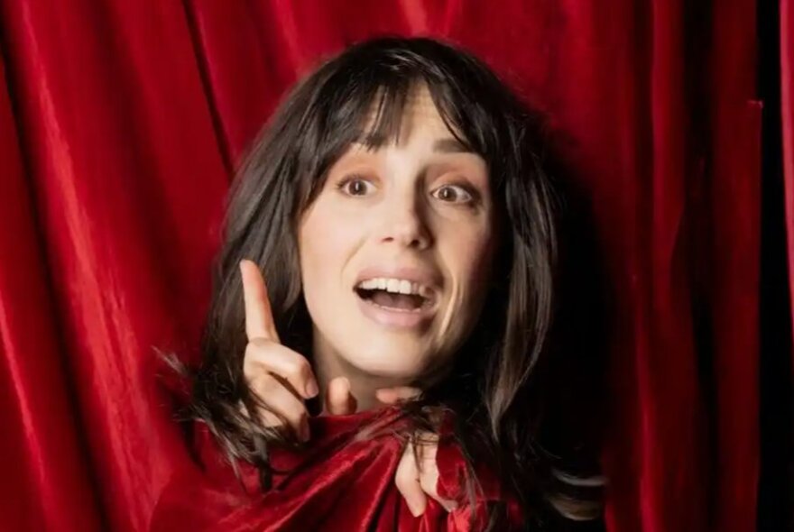Woman with dark hair, her head emerging from red curtains, holding her finger in the air.