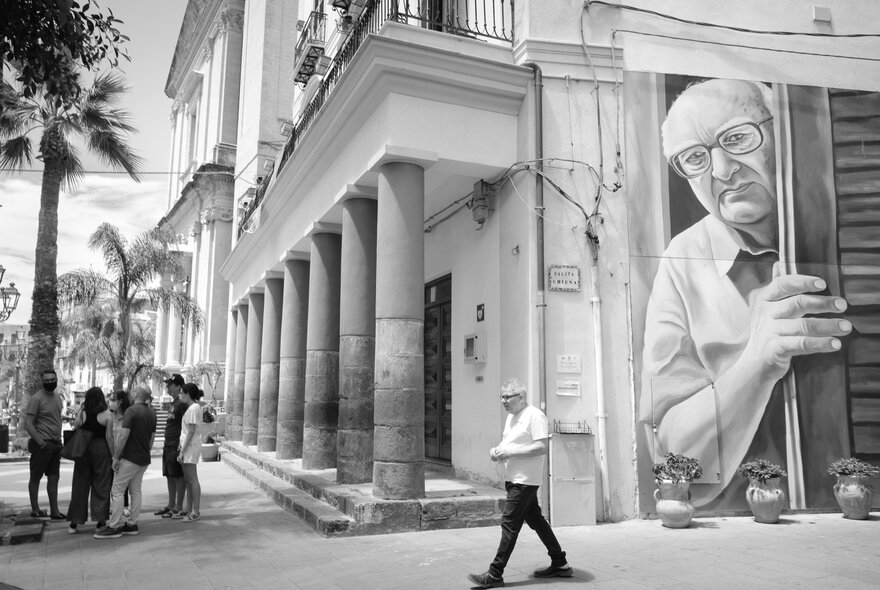Black and white image of a mural on the side of a building in Italy, with people in the street and tall palm trees.