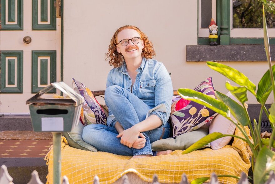 Comedian with curly red hair and glasses seated on a throw-covered sofa with cushions in a front garden.