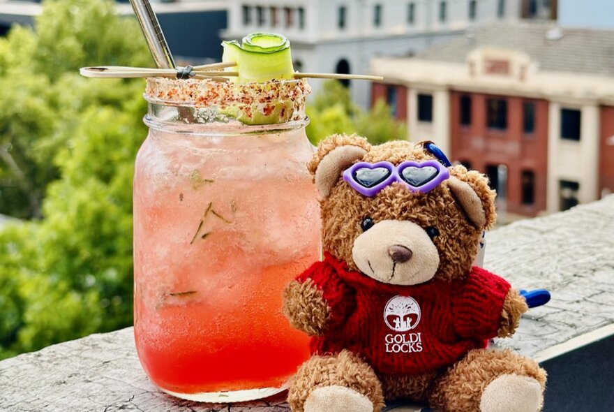 Toy bear and jamjar cocktail on a ledge overlooking buildings.