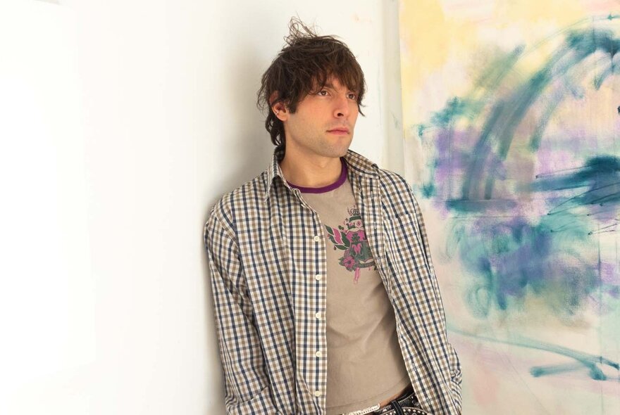 A young man with tousled brown hair,  casually leaning against a wall, wearing a check shirt over a t-shirt.
