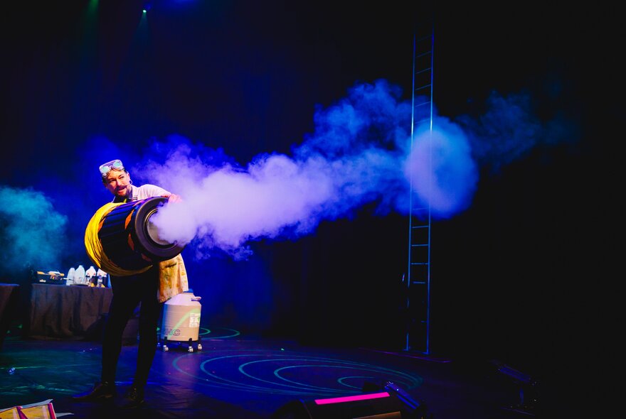 A darkened stage with a man shooting a large blue smoke ring from a large tube.