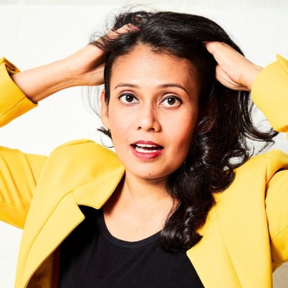 Comedian Kru Haralle in a yellow blazer with hands in her hair and mouth slightly open. 