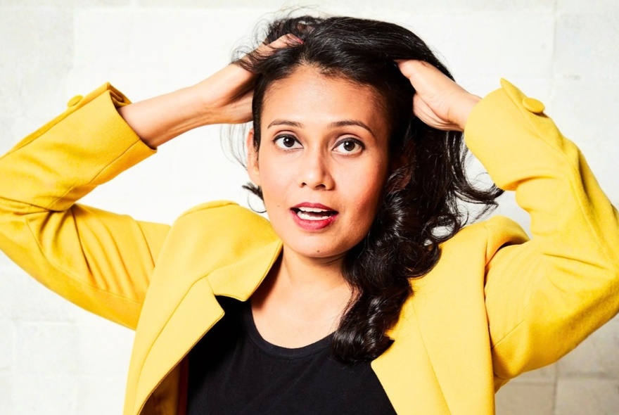 Comedian Kru Haralle in a yellow blazer with hands in her hair and mouth slightly open. 