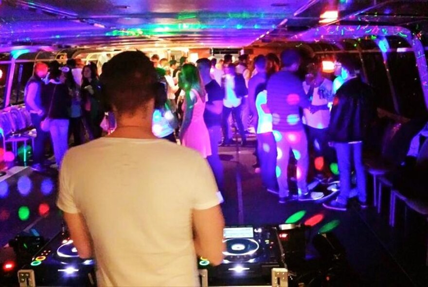 A DJ in a small party room with people mingling and coloured lights. 