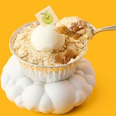A small cup of apple crumble and custard gelato with a silver spoon on an orangey-yellow background. 