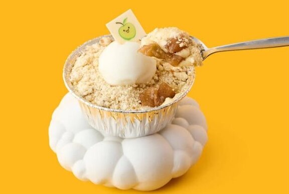A small cup of apple crumble and custard gelato with a silver spoon on an orangey-yellow background. 