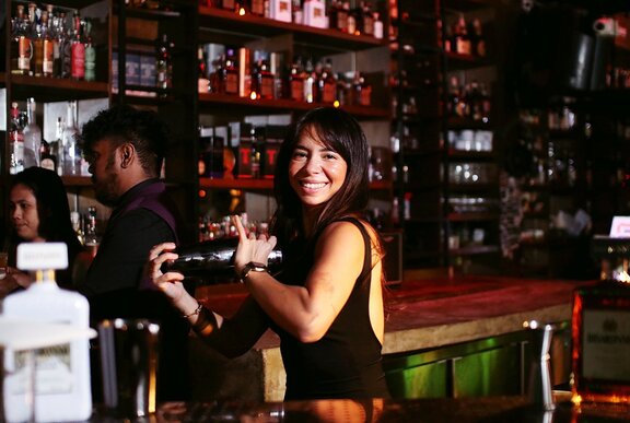 Woman shaking cocktails in a bar setting with bottles.