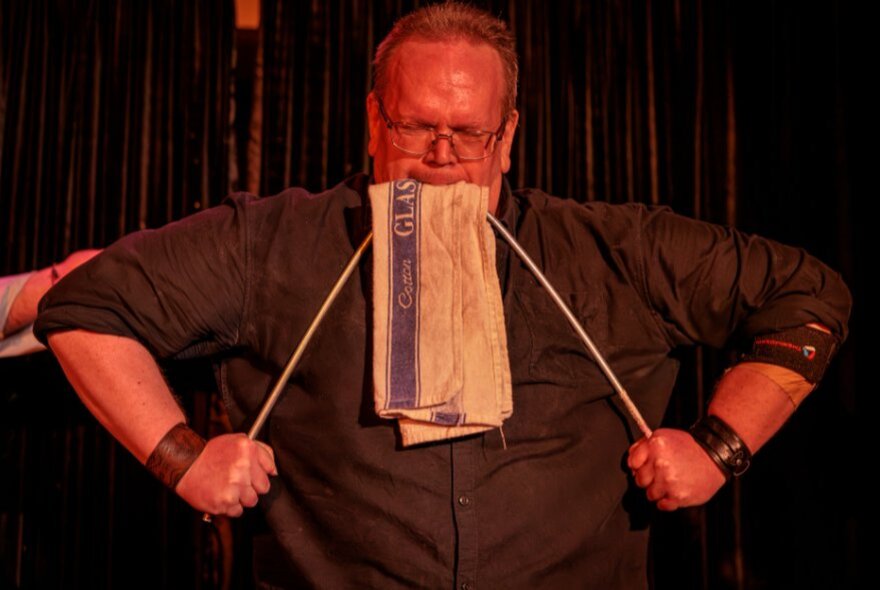 A man wearing glasses, with a tea towel hanging out of mouth, pulling on two white strings on either side of his mouth.