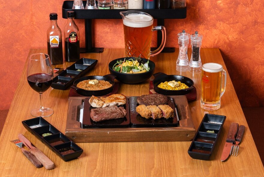 A table at Yappari Steak restaurant with hot plates of steak, several side dishes, glasses of beer and wine, plus cutlery and condiments.