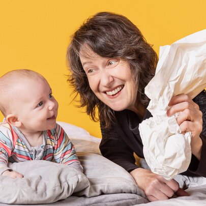 A smiling adult holding a scrunched-up bit of paper and interacting with a smiling infant lying on a cushioned surface, against a yellow background.