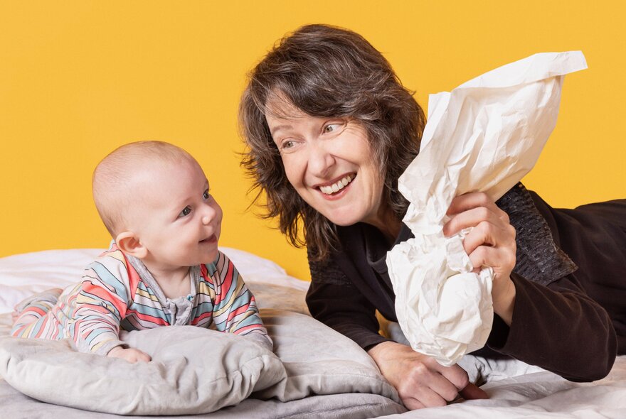 A smiling adult holding a scrunched-up bit of paper and interacting with a smiling infant lying on a cushioned surface, against a yellow background.