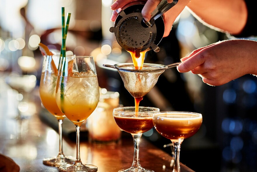 A bartender straining the contents of a cocktail shaker into two glasses with other cocktails nearby.