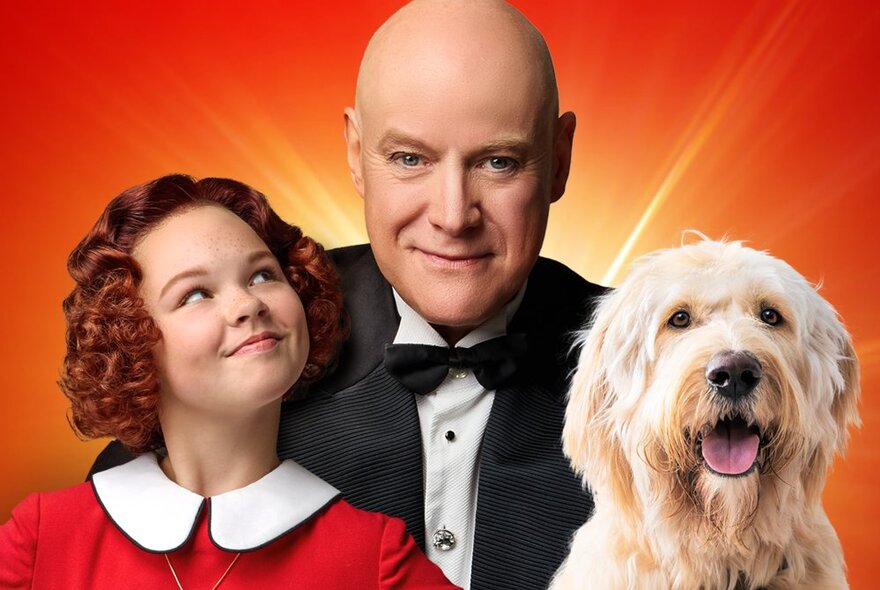 A poster for Annie the Musical with a grinning Annie, Daddy Warbucks and a scruffy dog with and orange-red background. 