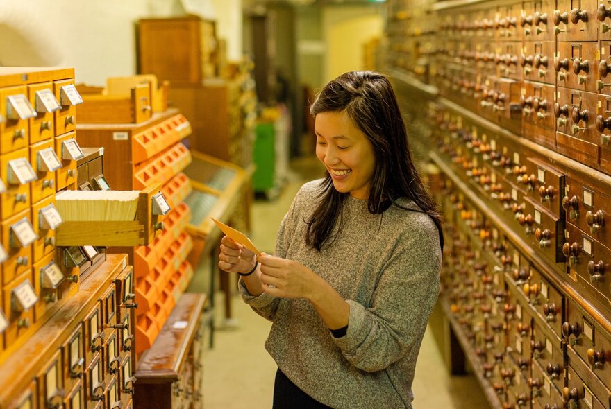 A person smiling while looking at paper library cards in a row of cataloguing shelves.