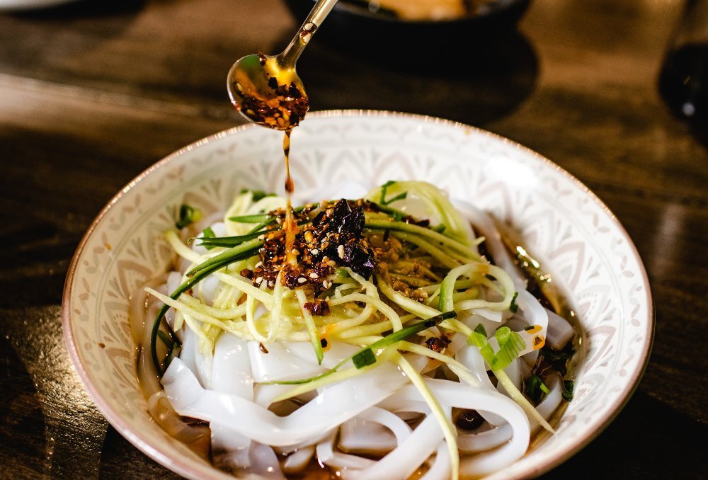 Person pouring a teaspoon of chilli oil over a bowl of thick white noodles with greens on top.