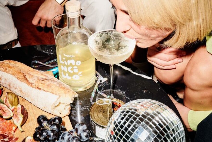 Looking down at a woman leaning forward to sip a cocktail, a disco ball and snacks nearby. 