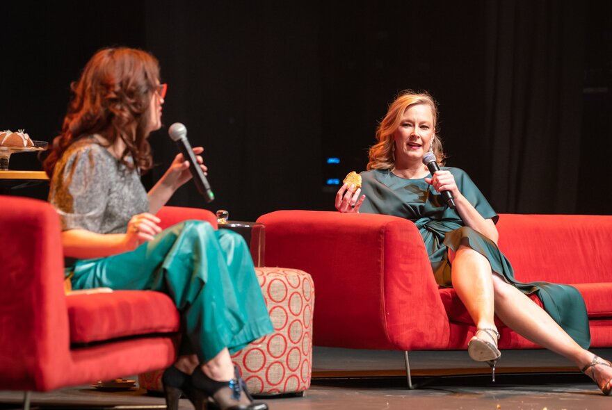 Annabel Crabb and Leigh Sales sitting on red couches on a theatre stage, both holding and talking into microphones. 