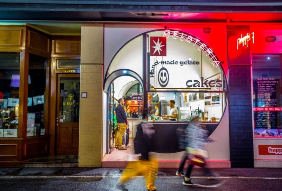 Pedestrians walking past a lit up gelato shop at night, the window is circular and there is a neon sign 