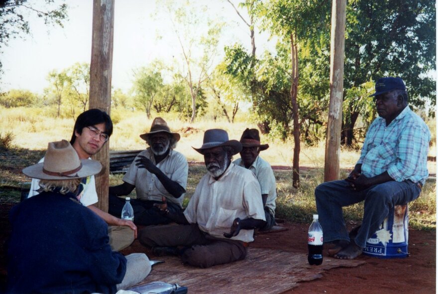 A Japanese man seated on the ground with a group of Aboriginal men under trees.