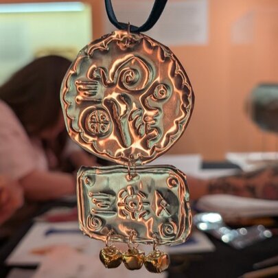 A gold-coloured metal pendant with a circular top section and a rectangular bottom section, featuring embossed abstract patterns, with blurred people working at a craft table in the background.