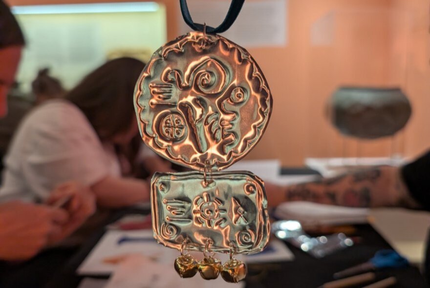 A gold-coloured metal pendant with a circular top section and a rectangular bottom section, featuring embossed abstract patterns, with blurred people working at a craft table in the background.