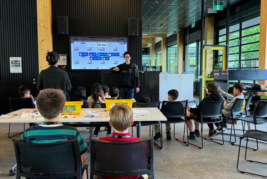 Kids in a casual classroom environment with a teacher up front and a digital board.