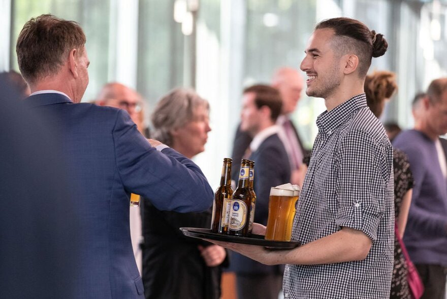 A waiter with a man-bun holding a tray of beers before the back of a man in a blue blazer. 