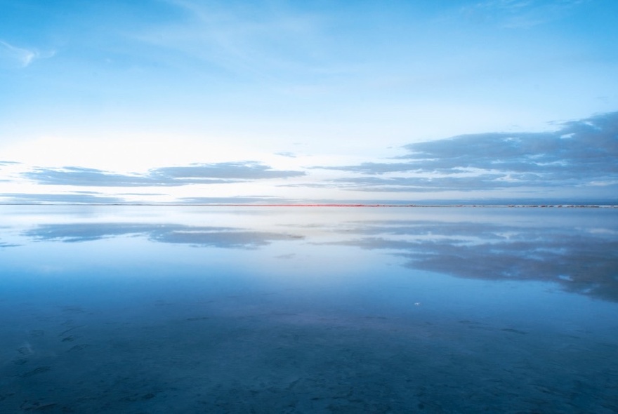 A vast, calm body of water reflecting a soft blue sky with thin, horizontal clouds.