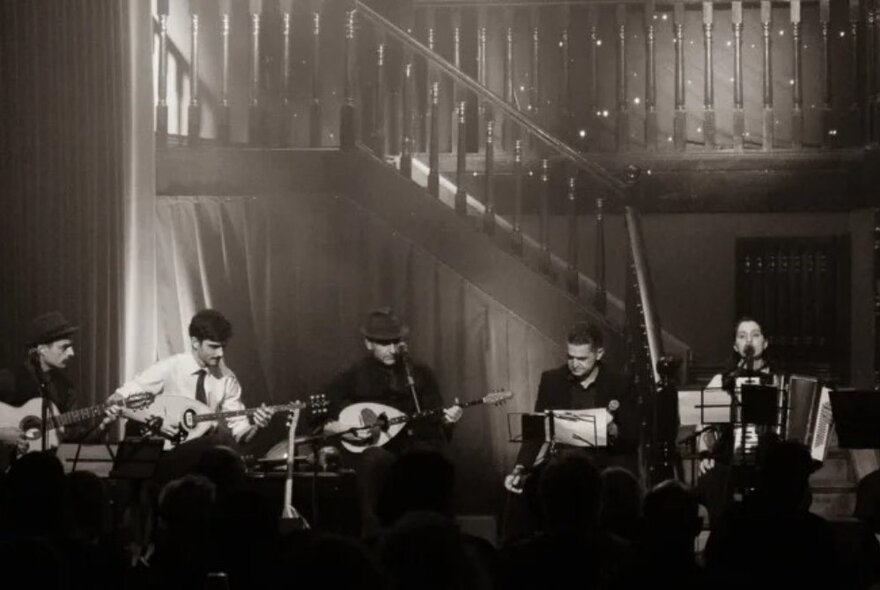 Musical ensemble seated at the foot of an internal staircase, playing ethnic instruments including the bouzouki.