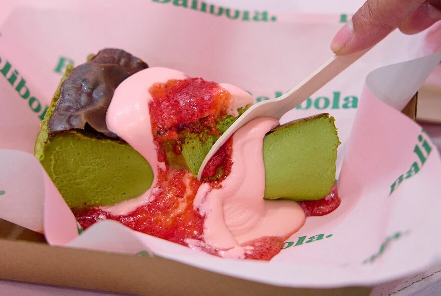 A person is cutting into a matcha cheesecake topped with strawberry sauce and jam.