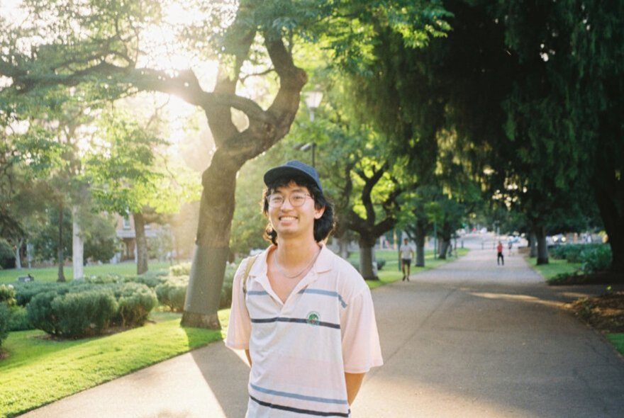 A man in a cap and open pink polo shirt smiles in a park on a pathway. 