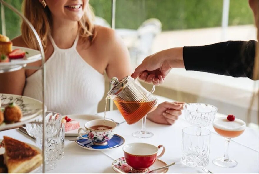 Hand pouring tea into an ornate teacup at a table set for afternoon tea while a woman wearing a tight white halterneck outfit smiles.