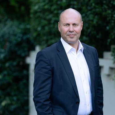 Bald man with blue jacket and white shirt, standing outside hedged house wall.