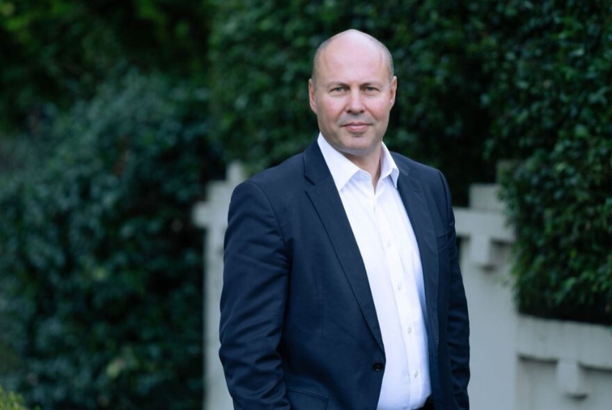 Bald man with blue jacket and white shirt, standing outside hedged house wall.