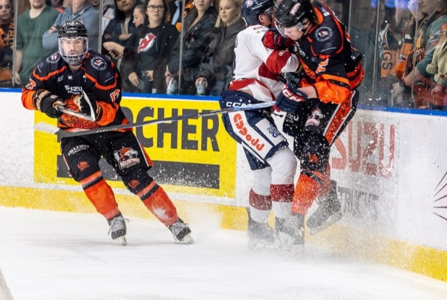 Two ice hockey players from opposing teams are smashed up against the barrier while a third skates past them.