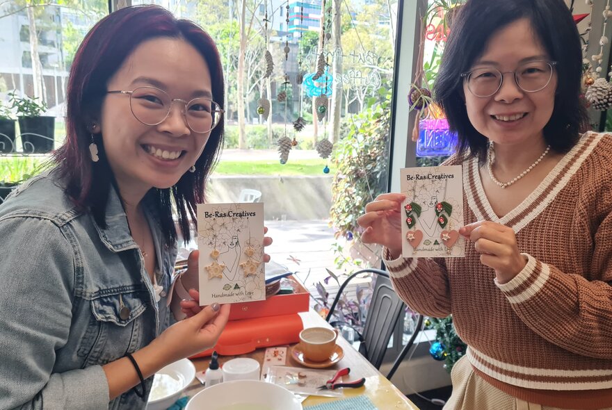 Two smiling workshop participants seated at a table with craft materials, holding up jewellery they have made.
