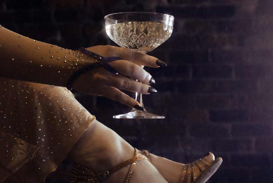 A woman's glitter-covered hand, holding a delicate crystal champagne glass in front of her lower legs.