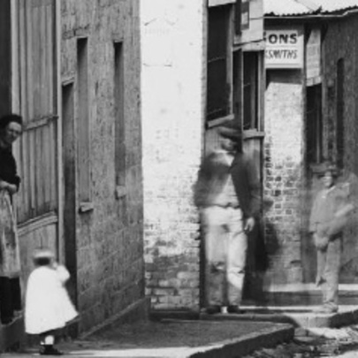 Historic black and white photograph shows Little Lonsdale Street with building facades and blurred figures walking along the street.