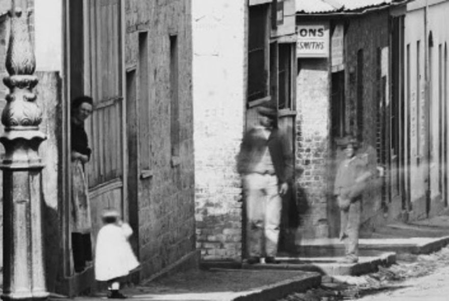 Historic black and white photograph shows Little Lonsdale Street with building facades and blurred figures walking along the street.
